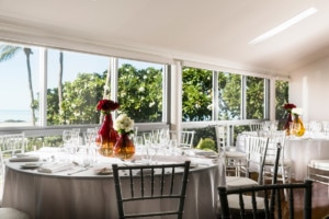 A light-filled wedding venue space is being decorated with round tables, chairs, dinnerware and wine glasses, and vases of roses.